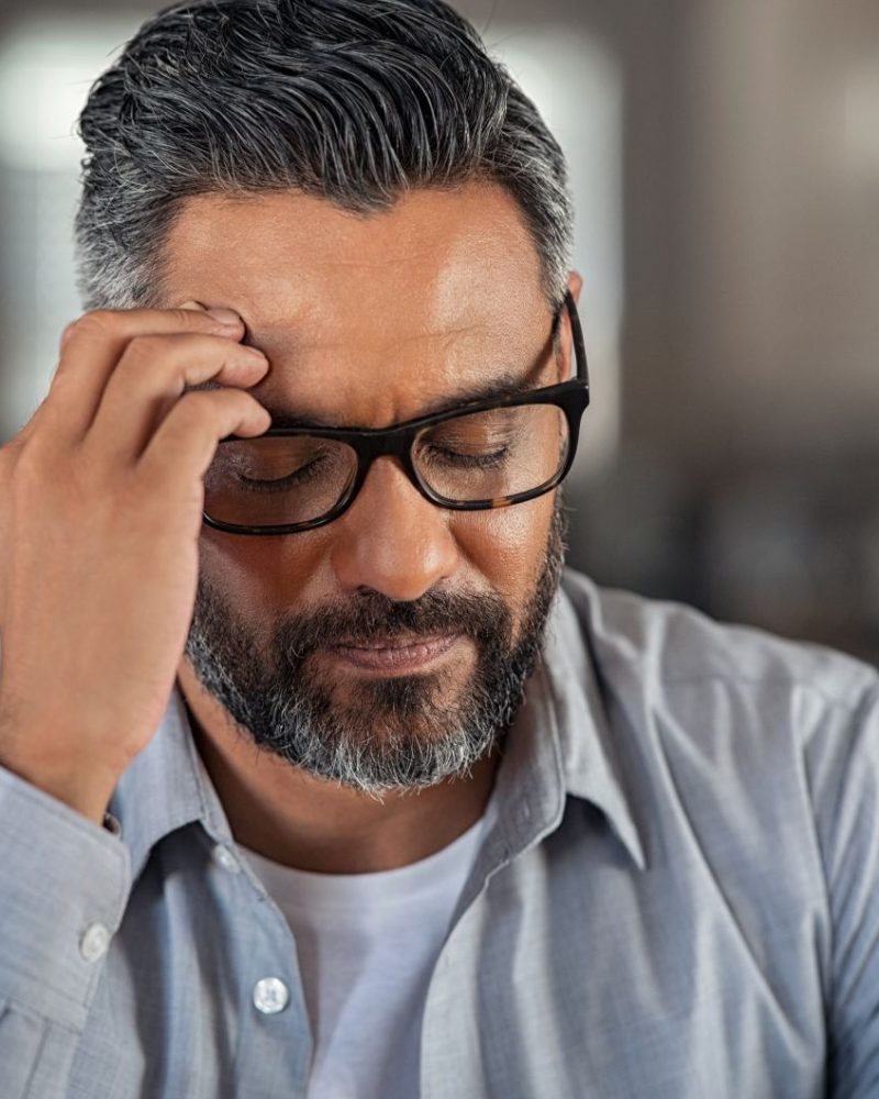 Frustrated middle aged man with hand on head sitting on couch at home. Close up face of stressed indian businessman wearing eyeglasses with eyes closed. Worried middle easter business man with terrible migraine, overload and overworked concept.